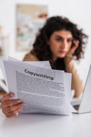 Blurred freelancer holding papers with copywriting lettering near laptop at home 