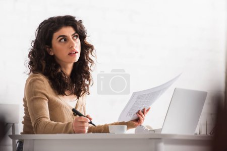 Rédacteur pensif travaillant avec des papiers et un ordinateur portable à la maison 