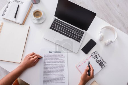Foto de Top view of copywriter writing on notebook near devices with blank screen and coffee on table - Imagen libre de derechos