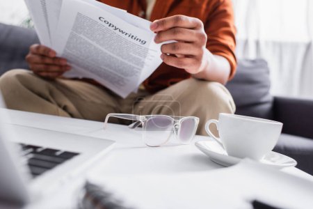 Foto de Cropped view of freelancer holding papers with copywriting lettering near coffee and eyeglasses at home - Imagen libre de derechos