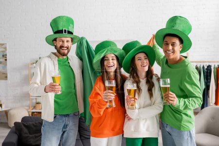 Foto de Cheerful and multicultural group of friends in green hats holding glasses of beer while celebrating Saint Patrick Day - Imagen libre de derechos