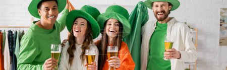 cheerful and multicultural group of friends in green hats holding glasses of beer while celebrating Saint Patrick Day, banner 