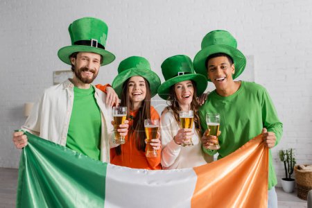 fröhliche und multikulturelle Freunde in grünen Hüten mit Biergläsern und irischer Flagge beim Feiern des St. Patrick Day 