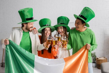 fröhliche und multikulturelle Freunde mit grünen Hüten, die Biergläser neben der irischen Flagge klappern, während sie den Saint Patrick Day feiern 