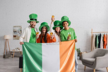 happy and multicultural friends in green hats holding glasses of beer and Irish flag while celebrating Saint Patrick Day 