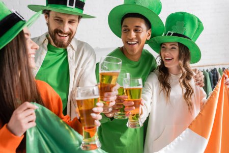 fröhliche und interrassische Freunde in grünen Hüten mit Biergläsern und irischer Flagge, während sie den Saint Patrick Day feiern 