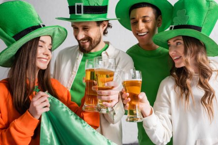 Foto de Cheerful and interracial friends in green hats clinking glasses of beer near Irish flag while celebrating Saint Patrick Day - Imagen libre de derechos