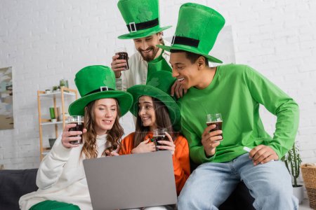 Foto de Positive and interracial friends in green hats holding glasses of dark beer while looking at laptop - Imagen libre de derechos