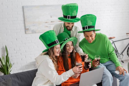 fröhliche und multiethnische Freunde mit grünen Hüten, die beim Blick auf den Laptop Gläser mit dunklem Bier klappern 