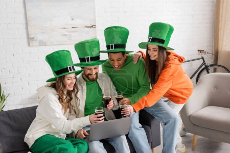 Foto de Happy and multiethnic friends in green hats holding glasses of dark beer during video call on laptop on Saint Patrick Day - Imagen libre de derechos