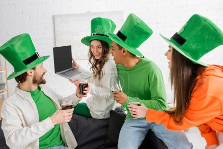 fröhliche Frau hält Laptop mit leerem Bildschirm neben multiethnischen Freunden mit Gläsern dunklem Bier am Saint Patrick Day
