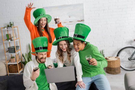 Téléchargez les photos : Cheerful woman holding laptop near multiethnic friends with glasses of dark beer during video call on Saint Patrick Day - en image libre de droit