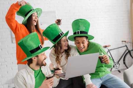 cheerful woman holding laptop near multiethnic friends with glasses of dark beer on Saint Patrick Day
