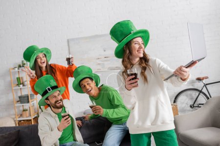 cheerful woman holding laptop near happy multiethnic friends with glasses of dark beer while celebrating Saint Patrick Day