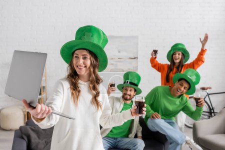 smiling woman holding laptop near happy multiethnic friends with glasses of dark beer while celebrating Saint Patrick Day