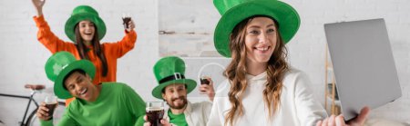 smiling woman holding laptop near happy multiethnic friends with glasses of dark beer while celebrating Saint Patrick Day, banner 