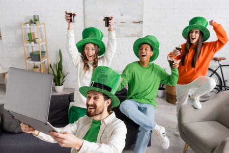 bearded man holding laptop near happy multiethnic friends with glasses of dark beer while celebrating Saint Patrick Day 