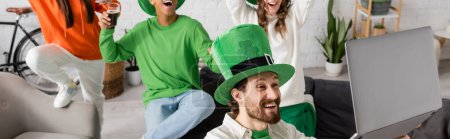 bearded man holding laptop near happy multiethnic friends with glasses of dark beer while celebrating Saint Patrick Day, banner 