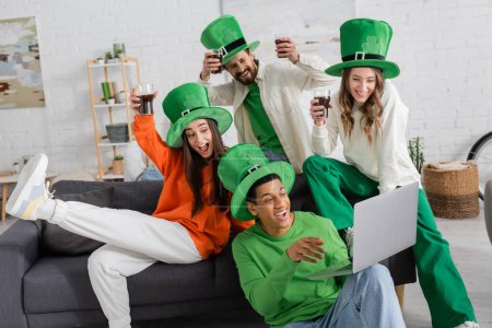happy african american man holding laptop during video call near cheerful friends toasting glasses of beer on Saint Patrick Day