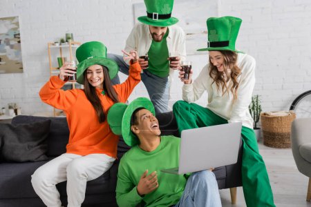 positive african american man holding laptop during video call near cheerful friends toasting glasses of beer on Saint Patrick Day