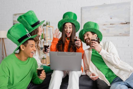 Aufgeregte Frau winkt bei Videoanruf in der Nähe von Freunden mit grünen Hüten am Saint Patrick Day