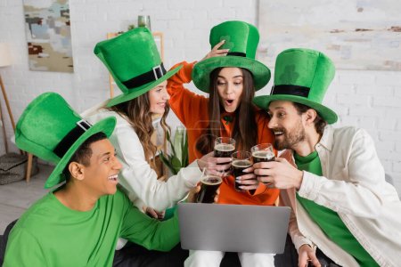 Foto de Excited woman looking at laptop during video call while toasting glasses of beer with interracial friends on Saint Patrick Day - Imagen libre de derechos