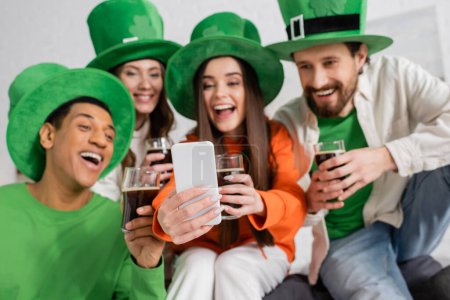 Photo for Happy interracial friends looking at smartphone during video call while holding glasses of dark beer on Saint Patrick Day - Royalty Free Image