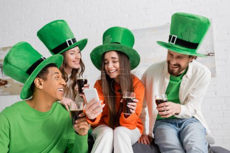 Foto de Happy interracial friends looking at friend with smartphone while holding glasses of dark beer on Saint Patrick Day - Imagen libre de derechos