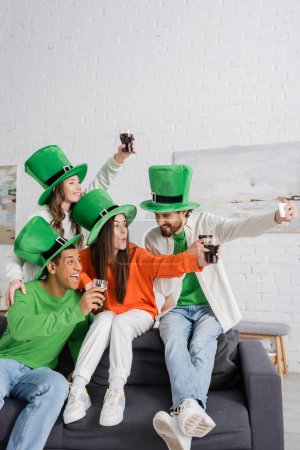 Foto de Alegre barbudo tomando selfie con felices amigos interracial sosteniendo vasos de cerveza oscura en el día de San Patricio - Imagen libre de derechos
