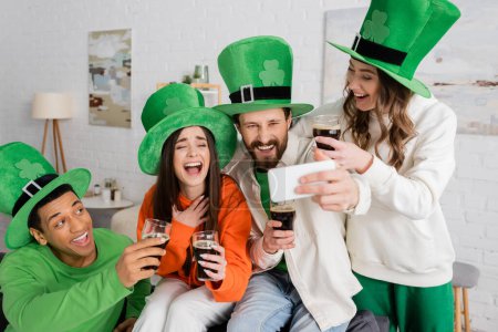 Téléchargez les photos : Homme barbu positif prenant selfie avec des amis interraciaux heureux tenant des verres de bière noire le jour de la Saint Patrick - en image libre de droit