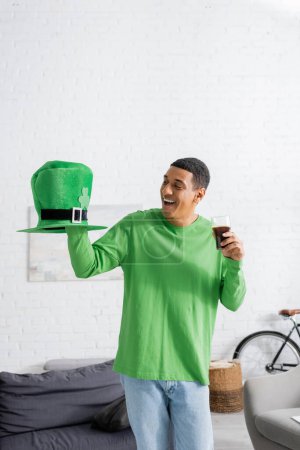 happy african american man holding glass of dark beer while looking at green hat on Saint Patrick Day