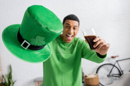 Foto de Hombre afroamericano feliz sosteniendo vaso de cerveza oscura y sombrero verde en el día de San Patricio - Imagen libre de derechos