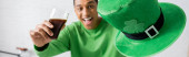 happy african american man holding glass of dark beer and green hat on Saint Patrick Day, banner  hoodie #639065884