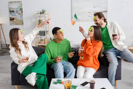 woman pointing at bearded man with sticky note on forehead holding Irish flag while playing guess who game with interracial friends 