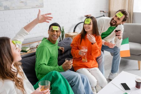 happy interracial friends with sticky notes on foreheads looking at woman while playing guess who game on Saint Patrick Day