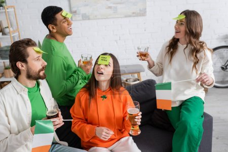 multiethnische Freunde mit klebrigen Zetteln auf der Stirn, die Alkoholgetränke in der Hand halten und erraten, wer am Saint Patrick Day spielt