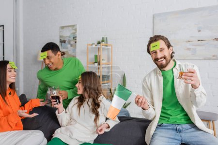 glücklicher bärtiger Mann mit Apfelwort auf klebrigem Zettel mit irischer Flagge und Getränk bei Freunden am Saint Patrick Day