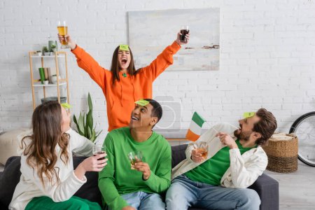 Mujer excitada sosteniendo cerveza cerca de amigos interracial con bandera irlandesa jugando quién soy en casa 