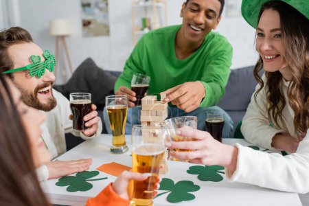 Cheerful women clinking alcohol drinks while interracial friends playing wood blocks game during saint patrick day 