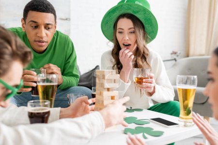 Pensive gemischtrassige Freunde mit Bier spielen Holzklötzchen-Spiel, während sie den Patrick-Tag zu Hause feiern 