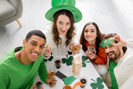 Overhead view of interracial friends celebrating saint patrick day and looking at camera at home 