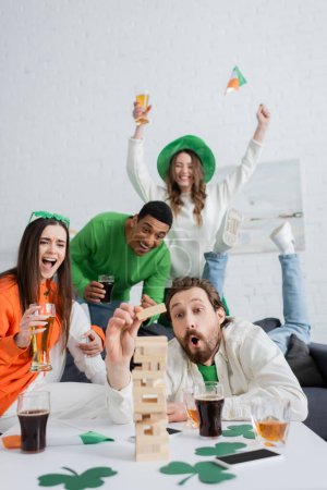 Excited interracial friends playing wood blocks game near beer and whiskey at home 