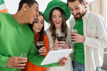 Afroamerikaner hält digitales Tablet in der Nähe von Freunden mit Bier während des Patronatsfestes zu Hause 
