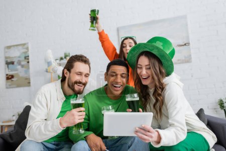 Foto de Mujer emocionada sosteniendo cerveza verde y tableta digital cerca de amigos multiétnicos en casa - Imagen libre de derechos