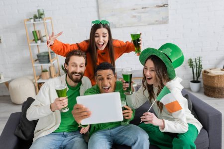 Aufgeregte multiethnische Freunde mit irischer Flagge und grünem Bier mit digitalem Tablet zu Hause 