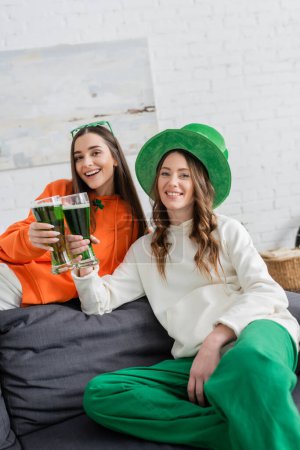 Smiling young women holding green beer and looking at camera at home 