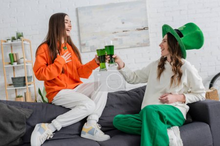 Cheerful young women clinking green beer and talking on couch on saint patrick day 