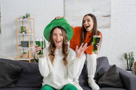 Smiling woman in green hat looking at camera near friend with beer at home 