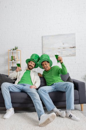 Positive multiethnic men in hats holding green beer during saint patrick celebration at home 