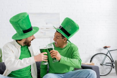 Emocionados amigos multiétnicos en sombreros verdes que tintinean cerveza durante el día de San Patricio en casa 